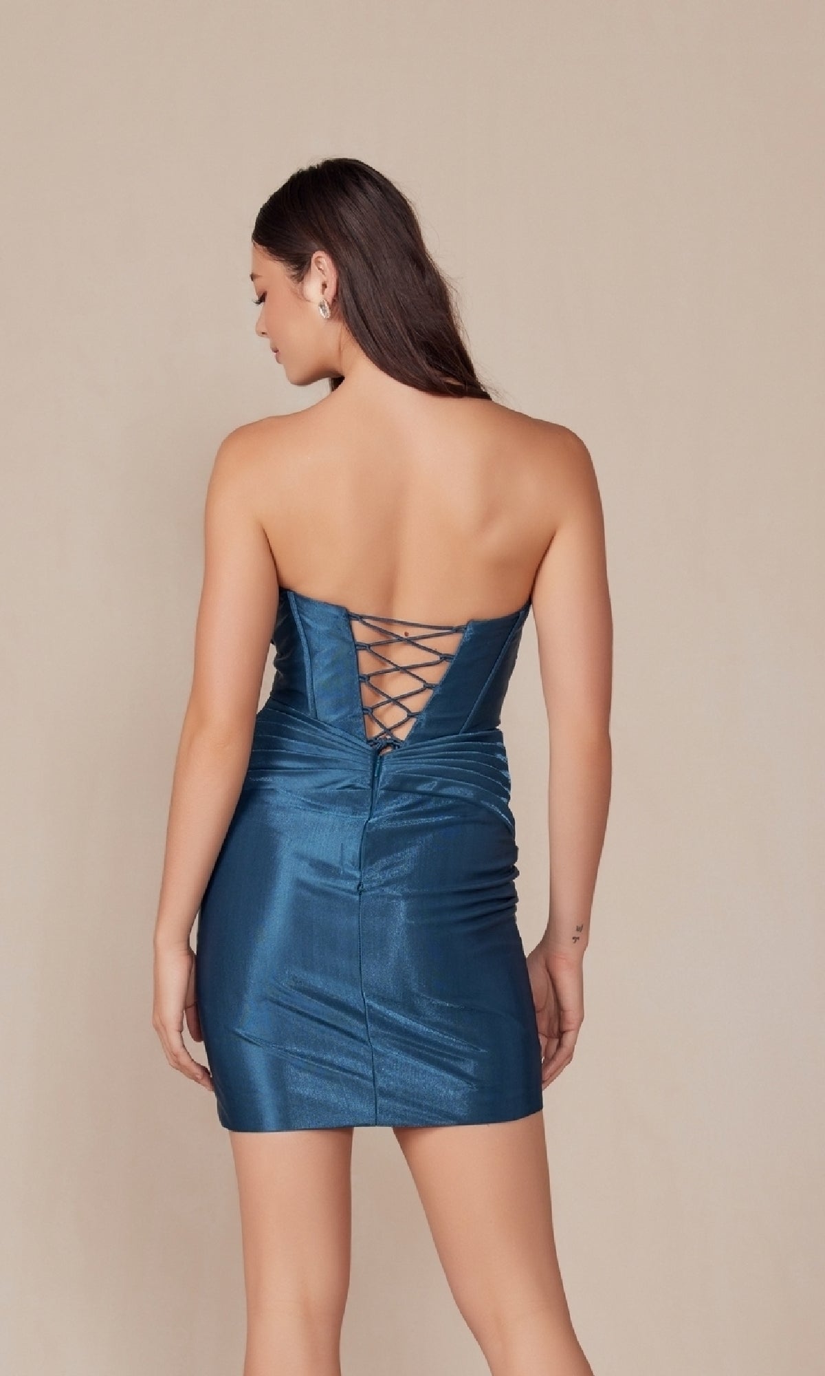 A woman faces away, wearing the Twist-Front Strapless Short Homecoming Dress T867 in shiny blue metallic fabric, featuring a lace-up back and corset-bodice mini silhouette, posed against a plain beige background.